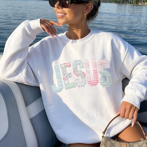 May include: White sweatshirt with the word "JESUS" in patchwork letters. The letters are in various pastel patterns, including floral, gingham, and polka dots. The sweatshirt is a crew neck style.