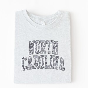 Może przedstawiać: Jasnoszara koszulka z napisem "NORTH CAROLINA" w szarym kamuflażu. Koszulka jest starannie złożona, eksponując nadrukowany wz&oacute;r.