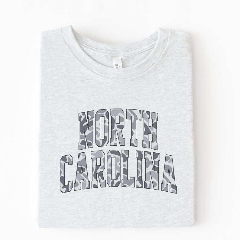 Może przedstawiać: Jasnoszara koszulka z napisem "NORTH CAROLINA" w szarym kamuflażu. Koszulka jest starannie złożona, eksponując nadrukowany wz&oacute;r.