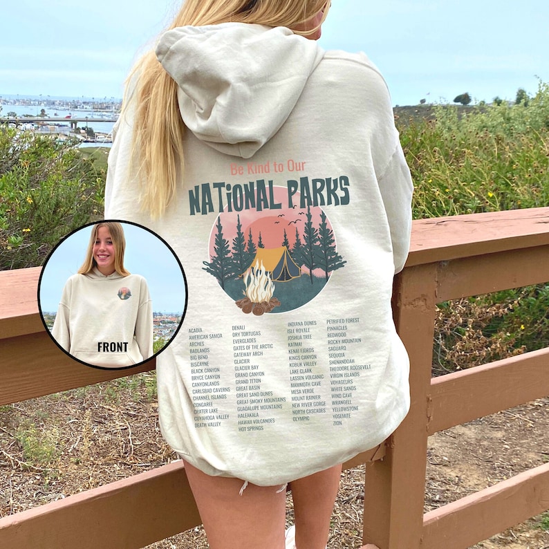 May include: A beige hoodie with a graphic print of a campfire scene in a forest. The text "Be Kind to Our National Parks" is printed above the graphic. A list of national parks is printed below the graphic.
