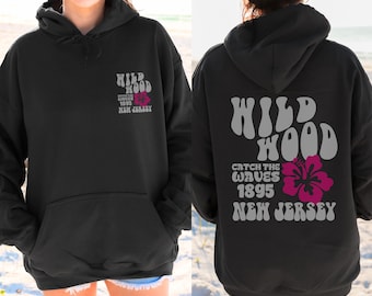 May include: Black hoodie with a front pocket and a hood. The front and back of the hoodie feature the text "WILD WOOD" and "CATCH THE WAVES 1895 NEW JERSEY" in a retro font, along with a pink hibiscus flower graphic.