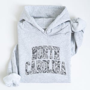 Może przedstawiać: Jasnoszara bluza z kapturem z napisem "NORTH CAROLINA" na piersi w kamuflażu. Bluza ma kaptur ze sznurkiem i długie rękawy. Tekst jest pogrubiony.