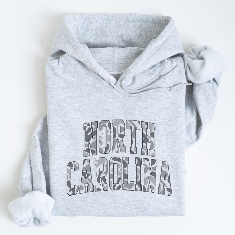 Może przedstawiać: Jasnoszara bluza z kapturem z napisem "NORTH CAROLINA" na piersi w kamuflażu. Bluza ma kaptur ze sznurkiem i długie rękawy. Tekst jest pogrubiony.
