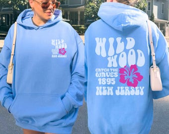 Könnte beinhalten: Hellblauer Kapuzenpullover mit Fronttasche und Kordelzugkapuze. Auf der Vorderseite des Hoodies steht der Text "Wild Wood" und eine rosa Blumengrafik. Auf der Rückseite des Hoodies steht der Text "Wild Wood, Catch the Waves 1895, New Jersey".