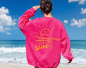 May include: A vibrant pink sweatshirt with the words "let your light shine" in yellow script and a sun graphic. The sweatshirt is a long-sleeved, crew neck style. The background shows a beach with blue sky and ocean.