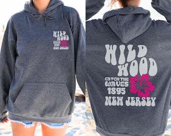 May include: A dark gray hoodie with a front pocket and drawstring hood. The front features the text "WILD WOOD" and "CATCH THE WAVES 1895 NEW JERSEY" with a pink flower graphic. The back of the hoodie has the same text and flower.