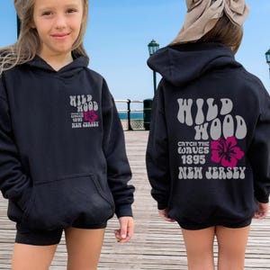 Może przedstawiać: Czarna bluza z kapturem z szarym i r&oacute;żowym nadrukiem graficznym z napisem "Wildwood Catch the Waves 1895 New Jersey".