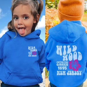 Może przedstawiać: Royal blue bluza z kapturem z białym nadrukiem graficznym, na kt&oacute;rym widnieje napis "Wildwood Catch the Waves 1895 New Jersey" z r&oacute;żowym kwiatem hibiskusa.
