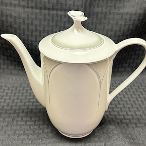 Puede incluir: Cafetera de cerámica blanca con tapa y ribete plateado. La cafetera tiene un cuerpo redondeado y un asa curva.
