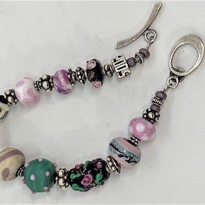 May include: A silver charm bracelet featuring an assortment of colourful glass beads, including pink, white, green, and black. The bracelet has a silver clasp and a charm that reads "SOUL".