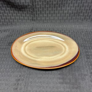 May include: An oval ceramic plate featuring a smooth, glossy finish with a gradient of beige and tan in the center, transitioning to a deep, dark brown rim. The surface has a subtle, reflective sheen and a clean, simple edge design.