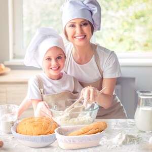 May include: Two people wearing white chef hats are baking bread. A round loaf of bread is in a white proofing basket and a long loaf of bread is in a rectangular proofing basket.