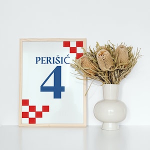 Puede incluir: Una impresi&oacute;n enmarcada con un patr&oacute;n de cuadros rojos y blancos y el texto "PERI&Scaron;IĆ 4" en azul. El marco es de madera clara y est&aacute; sobre una superficie blanca. Un ramo de flores secas en un jarr&oacute;n blanco est&aacute; a la derecha del marco.