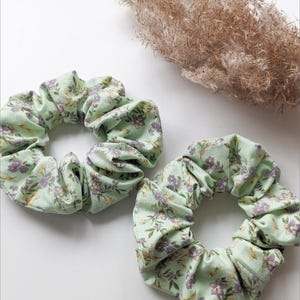 Puede incluir: Dos scrunchies verdes con estampado floral. Los scrunchies están hechos de tela y tienen un aspecto fruncido.