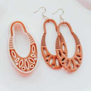 Op de afbeelding: Ovale oorbellen in terracotta-kleur met ingewikkelde uitgesneden patronen en goudkleurige oorhaken. Er is ook een bijpassende oorbelvormige koekjesvorm, met een witte omtrek en decoratieve uitsparingen. Op een wit oppervlak.