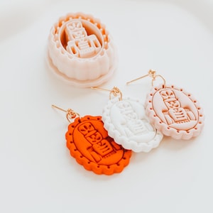 May include: Oval-shaped earrings in orange, white, and pink with a gold-colored post. Each earring features a detailed design with a central figure and decorative border. A matching cookie cutter is also present.