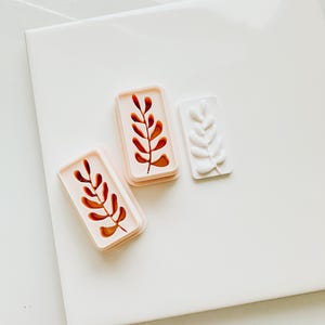 May include: Three rectangular silicone molds with leaf designs. Two molds are pink with brown leaf designs and one mold is white with a white leaf design.