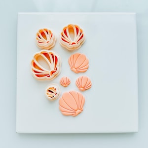 May include: A set of seven different sized and shaped cookie cutters in the form of seashells. The cutters are made of white plastic and are arranged on a white surface.