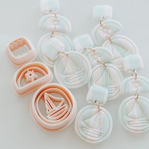 May include: Polymer clay earrings and clay cutters. The earrings have a sailboat design in a circular shape, with gold-coloured metal findings. The cutters are in various shapes, including a sailboat, a square, and a flower.
