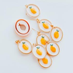 May include: A set of six lemon-shaped earrings with gold accents. The earrings are made of white clay with a yellow lemon design and green leaves. The earrings are displayed on a white surface with a silicone mould for making the earrings.