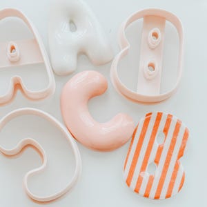 Op de afbeelding: Een verzameling lettervormige koekjesvormpjes en gegoten lettervormen. De set bevat een lichtroze A-vormpje, een witte A-vorm, een lichtroze B-vormpje, een lichtroze C-vorm, een lichtroze C-vormpje en een oranje-wit gestreepte B-vorm.