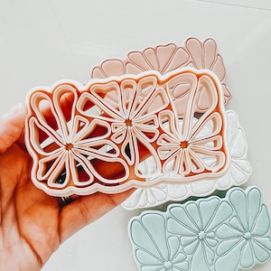 Puede incluir: Tres cortadores de galletas con forma de flores estilizadas. Los cortadores están hechos de plástico rosa claro, blanco y verde claro. Los cortadores están superpuestos.