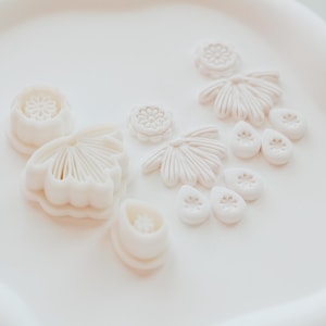 Może przedstawiać: Kolekcja białych foremek do ciastek i elementów dekoracyjnych w kształcie kwiatów, ułożonych na białej tacy. Kształty obejmują kwiaty, płatki i inne wzory botaniczne, idealne do pieczenia i rękodzieła.