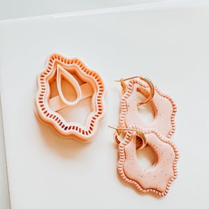 Puede incluir: Un cortador de pendientes de arcilla rosa claro y dos pendientes a juego con aros dorados. Los pendientes y el cortador tienen un borde festoneado y una textura moteada. El cortador tiene la forma de los pendientes.