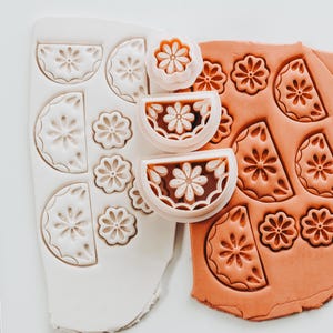 May include: A close-up of polymer clay and tools for creating floral designs. The image shows a white mold with flower shapes, a terracotta-colored clay with pressed flower designs, and flower-shaped cutters.