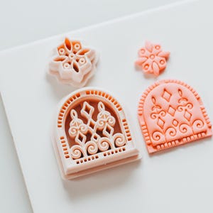 May include: Four light pink cookie cutters in various shapes, including a snowflake, a small flower, and two arched window designs with ornate scrollwork. The cutters are arranged on a white surface.