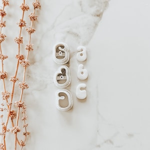 May include: White ceramic letter cookie cutters in the shapes of the letters a, b, c, d, and e. The cutters are arranged in two rows on a white marble surface.