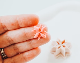 Può includere: Primo piano di due piccoli oggetti a forma di stella in una morbida tonalità rosa. Uno è tenuto in mano, mentre l'altro poggia su una superficie bianca. Gli oggetti hanno un design geometrico.