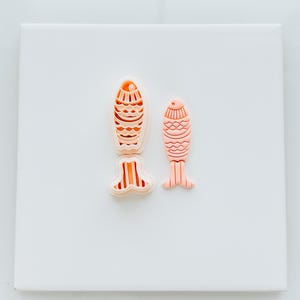 May include: Two pink fish-shaped cookie cutters, one larger than the other, on a white background. The larger cutter has a detailed design on the body and a striped tail.