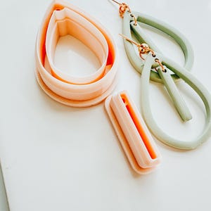 May include: A selection of craft tools and finished earrings. Peach-colored polymer clay cutters in teardrop shapes are next to sage green teardrop earrings with gold-tone findings. All items are on a white surface.