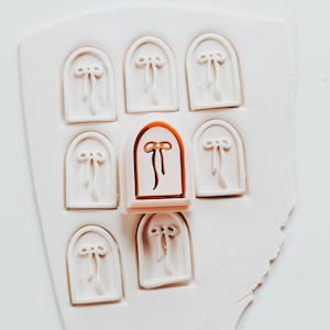 May include: A collection of white, arched polymer clay stamps with a bow design. One stamp is a copper color. The stamps are arranged on a white, irregular surface.
