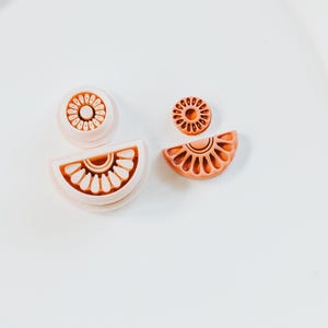 May include: Four light orange polymer clay cutters in the shape of a citrus fruit. The cutters include a circle, a half-circle, and a small circle, all with detailed interior designs. The cutters are on a white surface.