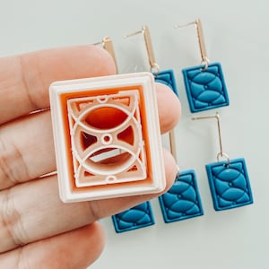 Puede incluir: Primer plano de unos pendientes y un molde a juego. Los pendientes tienen postes dorados y colgantes rectangulares azules con un diseño texturizado. El molde es de color rosa claro y naranja, con un patrón geométrico.