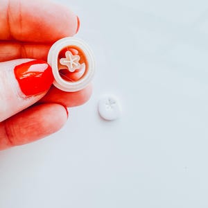 May include: A white plastic mold with a star-shaped design. The mold is being held in a hand with red nail polish.