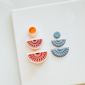 May include: A set of three polymer clay earrings, two are half-circles with intricate designs in red and white, and the other is a solid grey half-circle.
