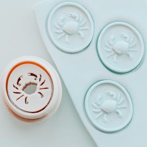 May include: A white and orange crab-shaped polymer clay cutter next to three light blue crab-shaped clay impressions. The cutter has a circular shape with a crab design cut out. The clay impressions are in a circular shape with a crab design.
