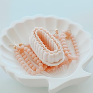 Könnte beinhalten: Eine weiße, muschelförmige Schale enthält ein kleines, weißes, geriffeltes Objekt und zwei Paar pfirsichfarbenen Perlenschmuck. Die Ohrringe und die Halskette bestehen aus kleinen, runden Perlen. Die Schale hat einen gewellten Rand.