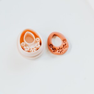 May include: Two terracotta-coloured Easter egg-shaped cookie cutters. One cutter is a complete egg shape with a floral design, and the other is a hollow egg shape with a floral design inside. The cutters are on a white surface.