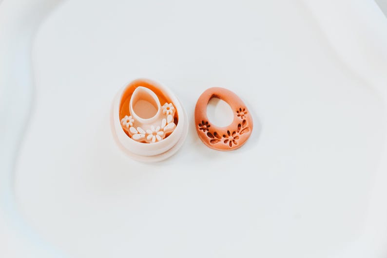 May include: Two terracotta-coloured Easter egg-shaped cookie cutters. One cutter is a complete egg shape with a floral design, and the other is a hollow egg shape with a floral design inside. The cutters are on a white surface.