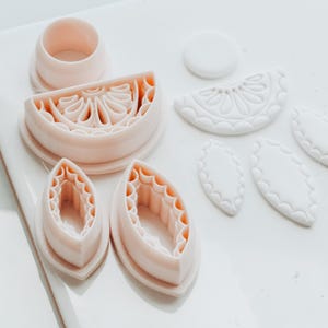 May include: A selection of light pink plastic cookie cutters in various shapes, including a citrus slice, leaves, and a circle. The cutters are arranged on a white surface, suggesting a baking or crafting theme.
