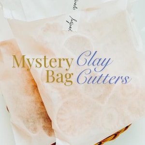May include: Two translucent white paper bags containing various shapes of clay cutters. The text Mystery Clay Cutters is printed in gold and blue on the front of the bags, with a small label at the top reading pottery grocery.
