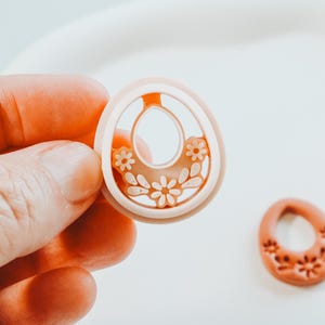 May include: A light peach-coloured earring component with a floral design is held in a hand. The earring has a central oval cutout and is decorated with small flowers. Another earring component with a floral design is next to it.