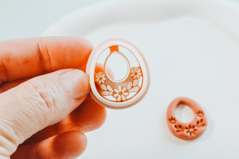 May include: A light peach-coloured earring component with a floral design is held in a hand. The earring has a central oval cutout and is decorated with small flowers. Another earring component with a floral design is next to it.