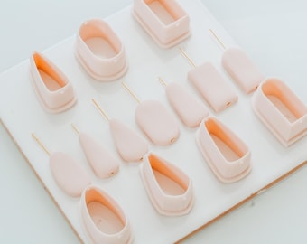May include: A collection of light pink polymer clay cutters and tools, including oval, rectangular, and teardrop shapes, arranged on a white surface. Each cutter has a gold-colored wire attached.
