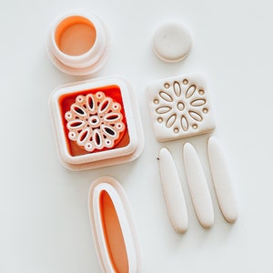 May include: A set of pink polymer clay molds for crafting. The molds include a square with a floral design, a circle, a rectangle, and three long, thin shapes.