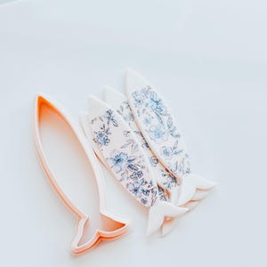 Op de afbeelding: Een set van vier witte koekjesvormen in de vorm van een vis met een bloemenpatroon. De vormen zijn in een rij gerangschikt met de staarten die elkaar overlappen. Een grotere, effen witte koekjesvorm in de vorm van een vis is ook inbegrepen.